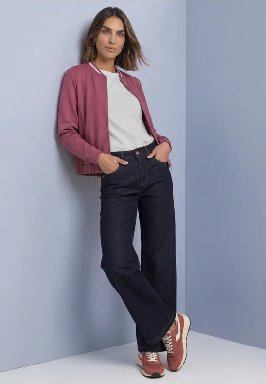 Frau in dunkelblauen, weiten Jeans, weißem T-Shirt und rosafarbenem Cardigan, lässig vor hellem Hintergrund.