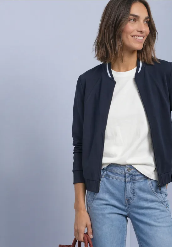 Une femme porte un t-shirt blanc, une veste bleue et un jean, tenant un sac rouge devant un mur bleu clair.