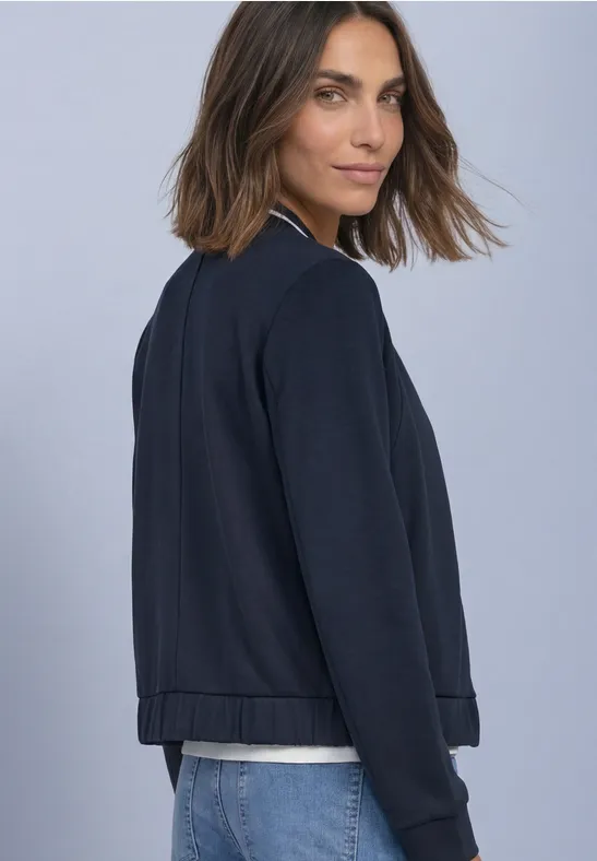 Femme portant une veste bleu marine, vue de côté, regard par-dessus l’épaule, fond clair.