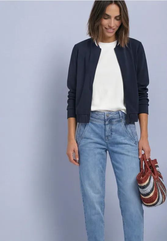 Femme porte un T-shirt blanc, un gilet bleu marine et un jean clair, elle tient un sac rayé coloré.