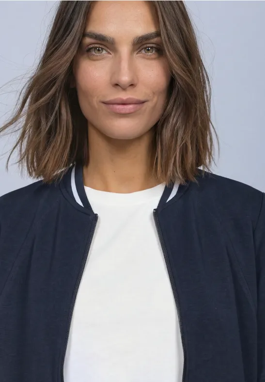 Une femme aux cheveux bruns mi-longs porte un t-shirt blanc et une veste sombre devant un fond clair.
