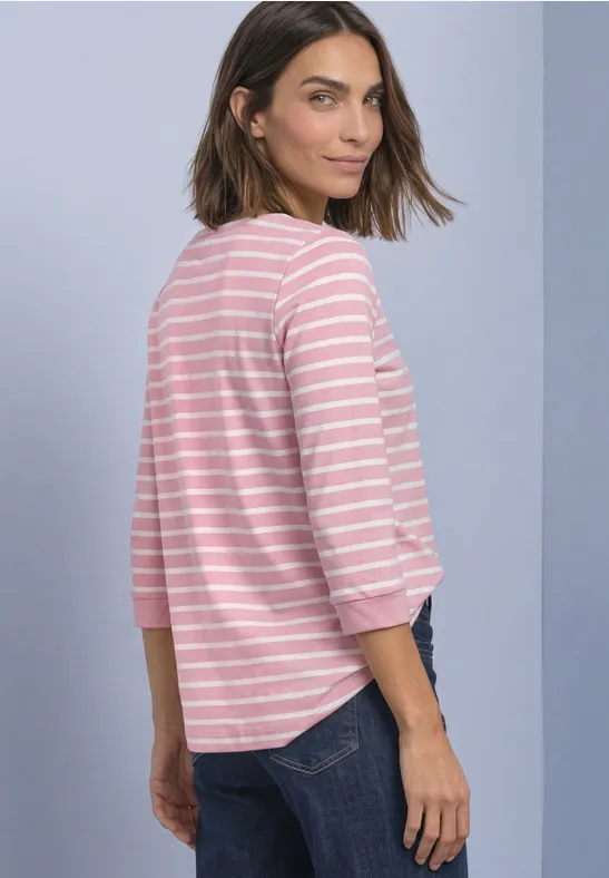 Frau steht seitlich, blickt über die Schulter, trägt rosa-weiß gestreiftes Shirt und Jeans vor neutralem Hintergrund.