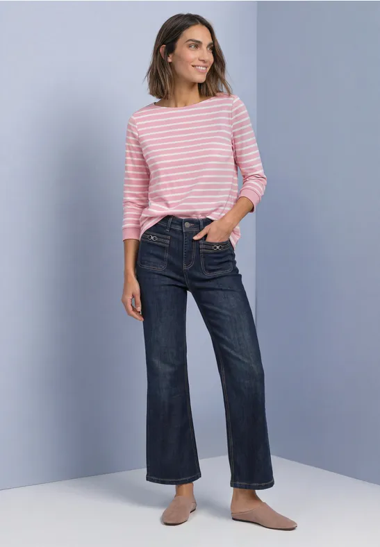 Frau mit rosa-weiß gestreiftem Pullover, weiten Jeans und Slippern vor neutralem Hintergrund.