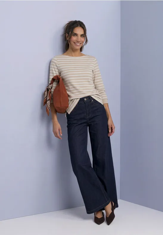 Frau lehnt an hellblauer Wand, trägt gestreiftes Top, weite Jeans, braune Schuhe und hält eine Leder-Tasche.