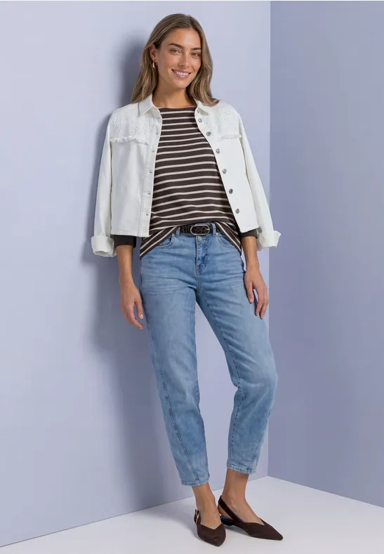 Frau in weißer Jeansjacke, gestreiftem Shirt, blauen Jeans und braunen Slippern vor hellem Hintergrund.