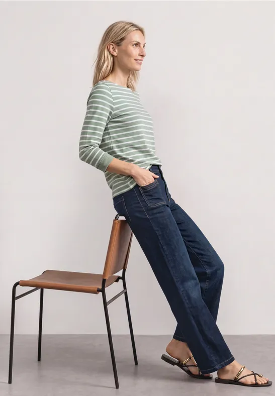 Frau in gestreiftem Langarmshirt, dunkelblauen Jeans und Sandalen lehnt elegant an einem Stuhl.