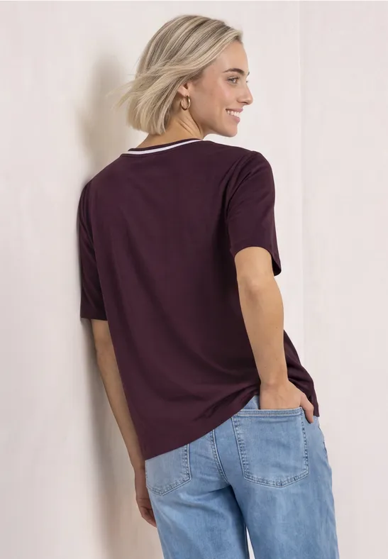 Junge Frau von hinten in lockerem lila T-Shirt mit weißem Kragen und hellblauen Jeans, lässig an Wand gelehnt.