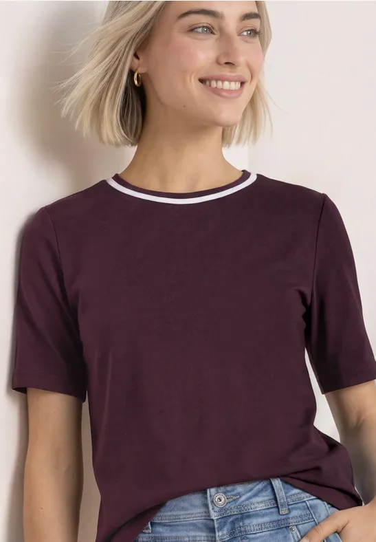 Frau mit blonden Haaren trägt burgunderrotes T-Shirt mit gestreiftem Kragen und Jeans, lächelt.