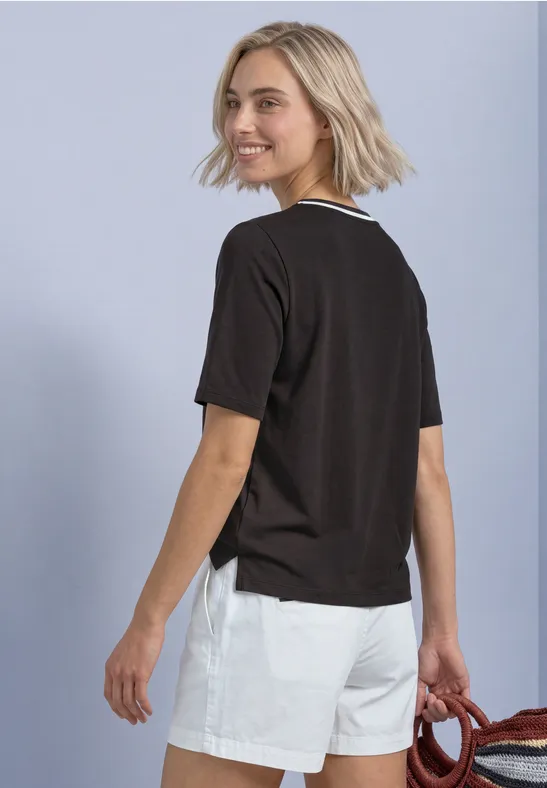Junge Frau mit blondem Bob, schwarzem T-Shirt und weißen Shorts, blickt über die Schulter, trägt rotes Accessoire