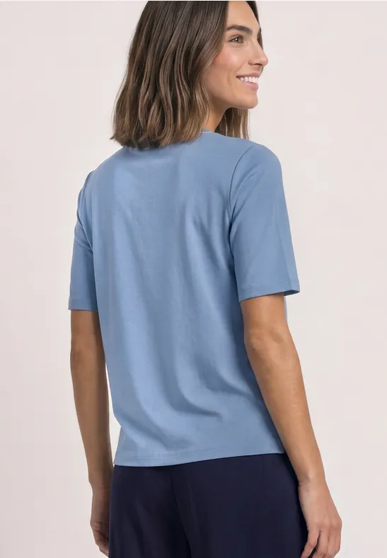 Frau trägt hellblaues T-Shirt und dunkle Hose, lächelt seitlich vor hellem Hintergrund.