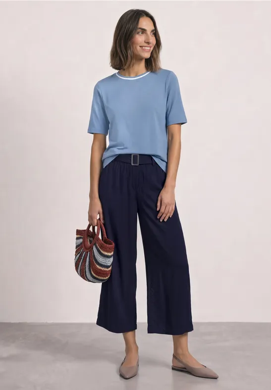Lächelnde Frau in blauem T-Shirt, weiter dunkelblauer Hose, trägt gestreifte Tasche und flache Schuhe.