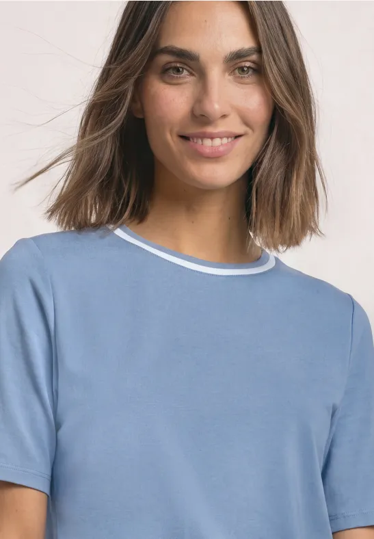 Eine lächelnde Frau mit braunen Haaren trägt ein hellblaues Oberteil vor hellem Hintergrund.