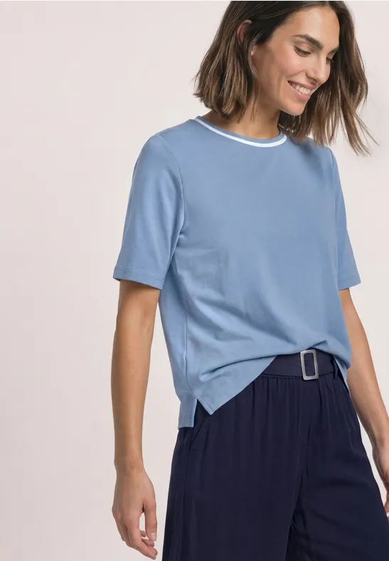 Eine lächelnde Frau in hellblauem T-Shirt mit weißem Kragen und weiten, dunkelblauen Hosen mit Gürtel.