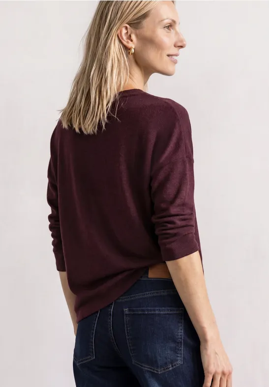 Eine Frau mit hellem Haar in weinrotem Pullover und Jeans steht seitlich vor weißem Hintergrund.