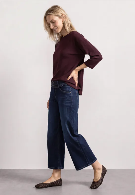 Frau in bordeauxrotem Oberteil, weiten blauen Jeans und braunen flachen Schuhen.