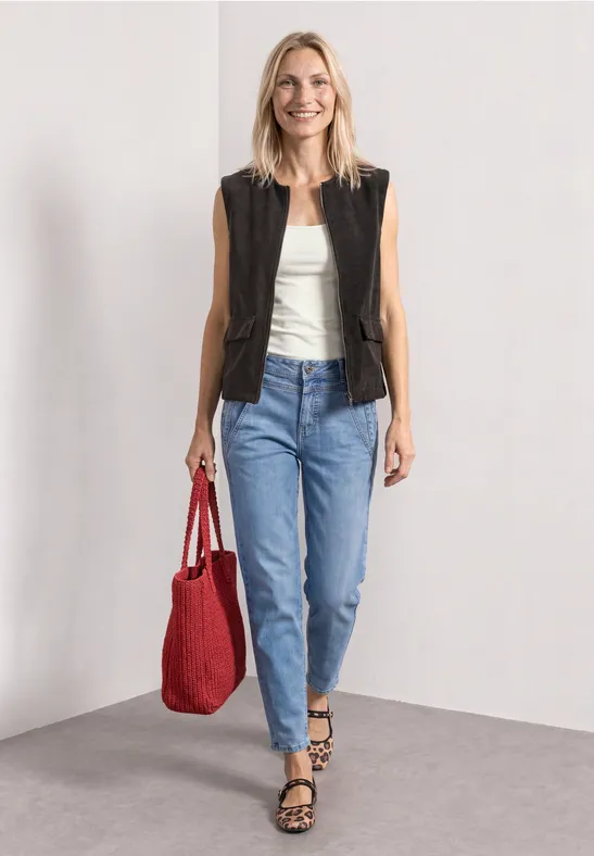 Een vrouw in jeans, wit topje en zwart vest draagt een rode tas en loopt met een glimlach.