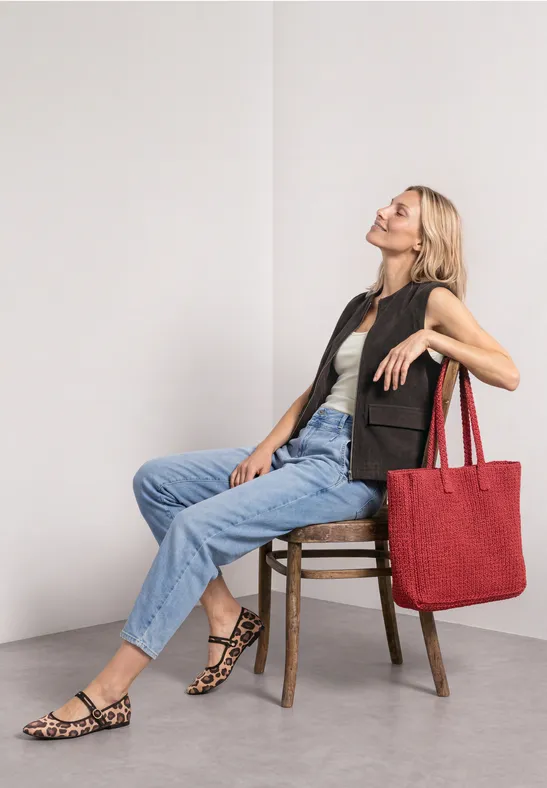 Vrouw zit ontspannen op een stoel voor een witte muur, draagt jeans, vest, sandalen en grote rode tas.