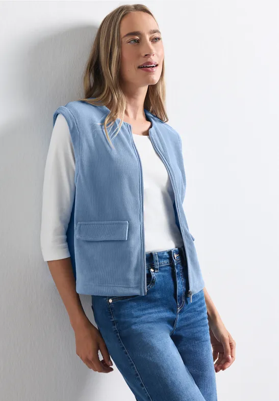 Femme avec chemise blanche, veste sans manches bleue et jean appuyée sur un mur blanc