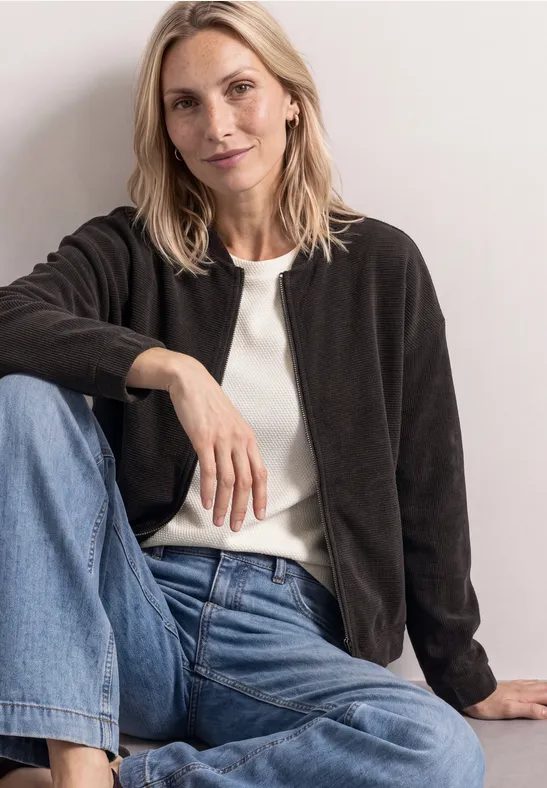 Eine entspannte Frau sitzt lässig in schwarzer Jacke, weißem Top und blauen Jeans.