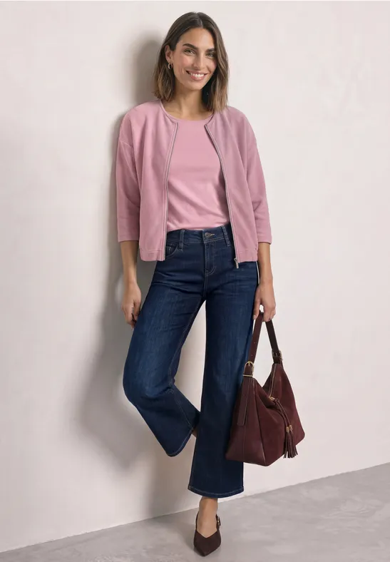 Eine lächelnde Frau lehnt an der Wand, trägt rosa Oberteile, dunkelblaue Jeans und hält eine dunkelrote Tasche.