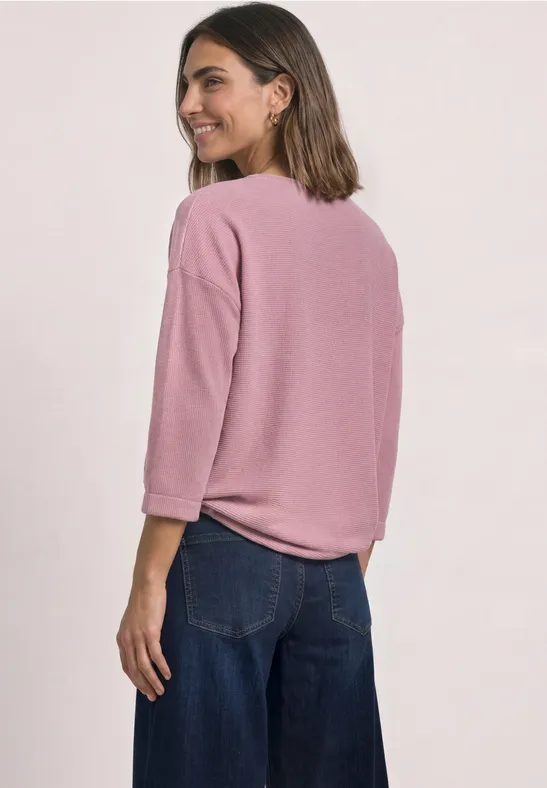 Eine Frau mit rosa Dreiviertelarmshirt und weiten Jeanshosen schaut über die Schulter und lächelt.