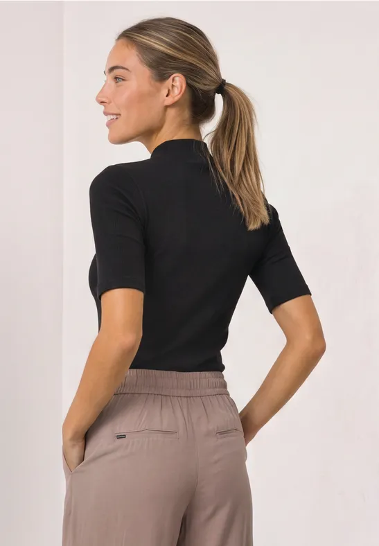 Frau im Profil mit Pferdeschwanz, schwarzes Kurzarmshirt und beigefarbene Bundfaltenhose, Hände in den Taschen.