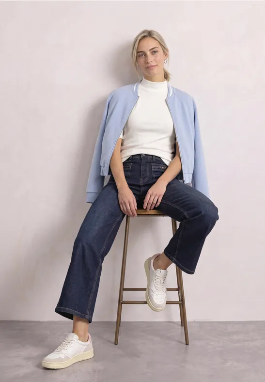 Frau sitzt lässig auf Hocker, trägt weiße Sneaker, weite Jeans, weißen Rollkragenpullover und hellblauen Cardigan.