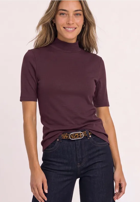 Eine lächelnde Frau trägt ein weinrotes Kurzarm-Longsleeve mit Stehkragen und Jeans mit Gürtel.