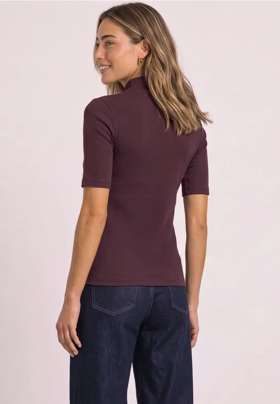 Frau von hinten, trägt ein bordeauxfarbenes Kurzarmshirt und dunkle Jeans.
