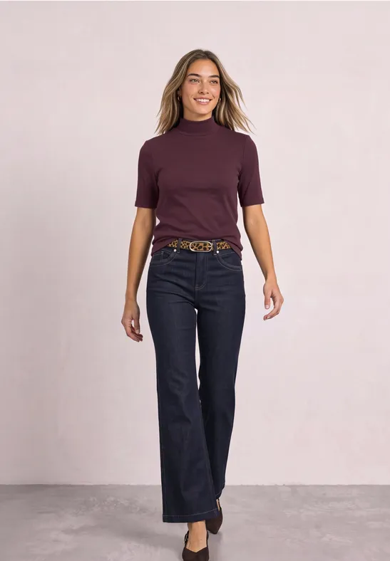 Eine Frau im lila Kurzarmshirt und dunkler Jeans läuft selbstbewusst auf neutralem Hintergrund.