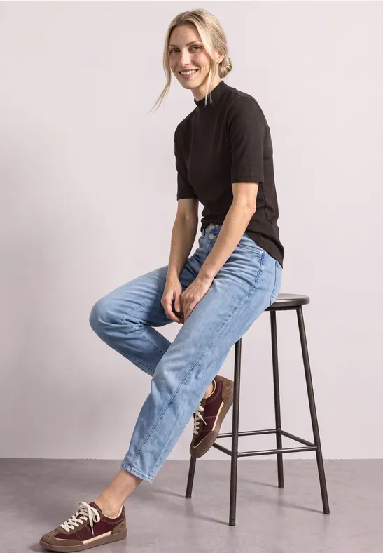 Frau sitzt lässig auf einem Hocker, trägt T-Shirt, Jeans und Sneakers und lächelt.