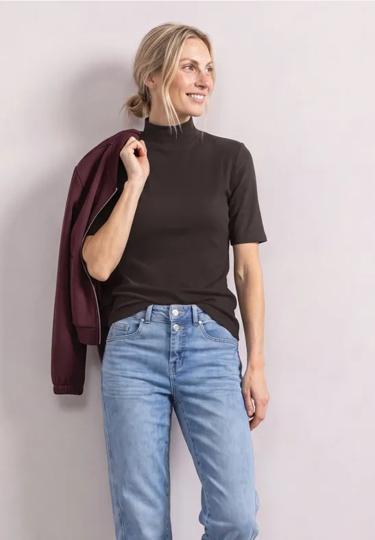 Frau in dunklem Kurzarmshirt, Jeans und weinroter Jacke über der Schulter, lächelnd vor neutralem Hintergrund.