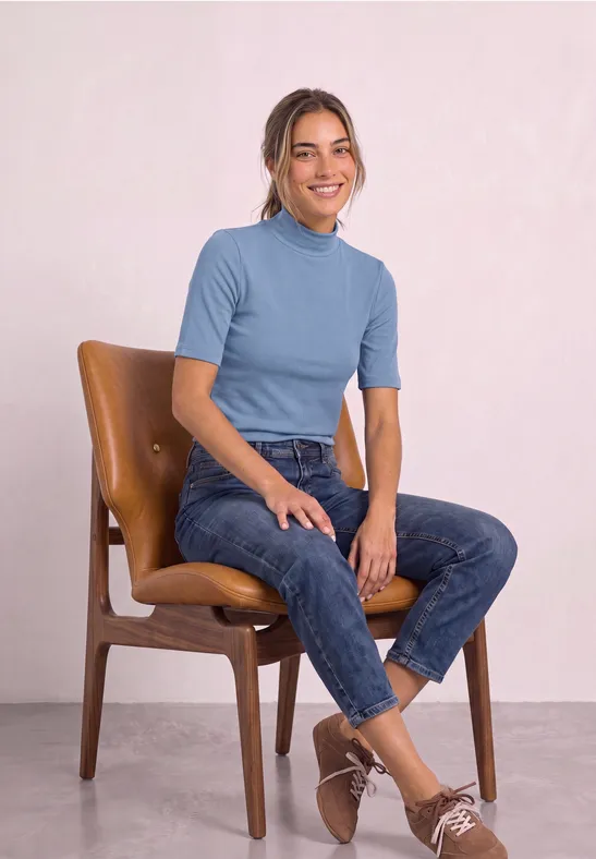 Junge Frau in hellblauem T-Shirt und Jeans sitzt lächelnd auf einem Holzstuhl.