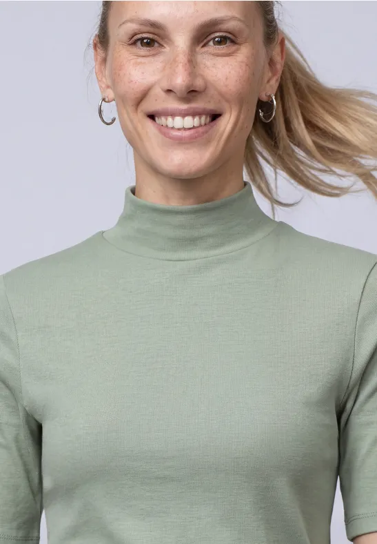 Lächelnde Frau mit Zopf, Creolen und grünem Kurzarm-Rollkragenpullover vor hellem Hintergrund.