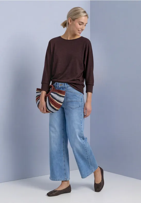 Frau mit blondem Dutt trägt dunkelroten Pullover, hellblaue weite Jeans, braune Ballerinas und gestreifte Tasche vor schlichtem Hintergrund.