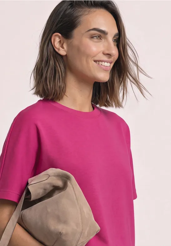 Vrouw lacht opzij, draagt een felroze T-shirt en houdt een beige tas vast.