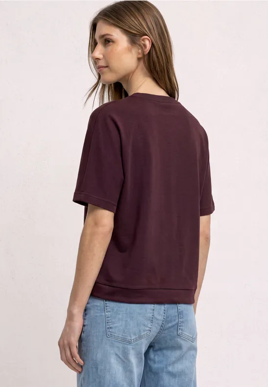 Jeune femme vue de dos et de profil, portant un t-shirt bordeaux et un jean bleu clair sur fond clair.