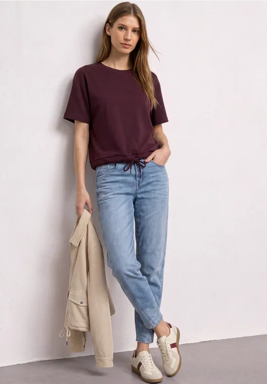 Jeune femme appuyée contre un mur blanc, en t-shirt bordeaux, jean bleu clair, baskets blanches et veste beige.
