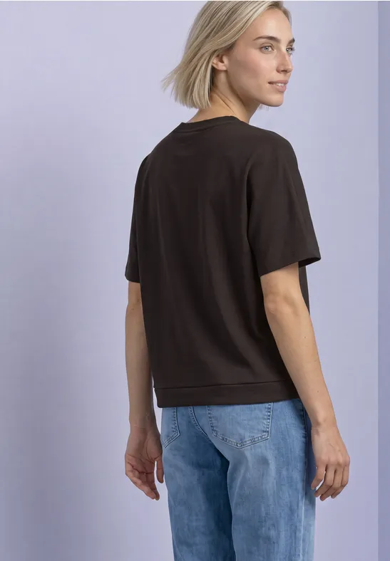 Blonde Frau in schwarzem T-Shirt und blauen Jeans, steht seitlich und blickt über die Schulter vor hellem Hintergrund.