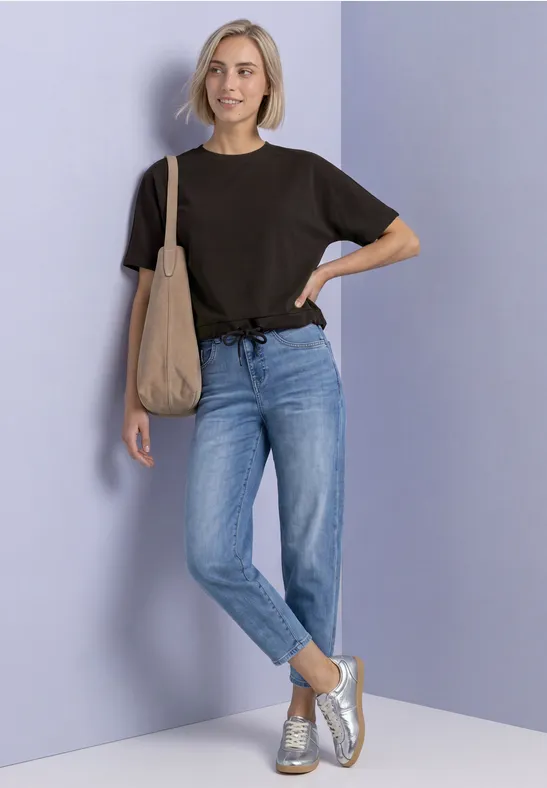 Frau in schwarzem T-Shirt, blauen Jeans und Sneakers, mit Stofftasche vor lila Hintergrund.