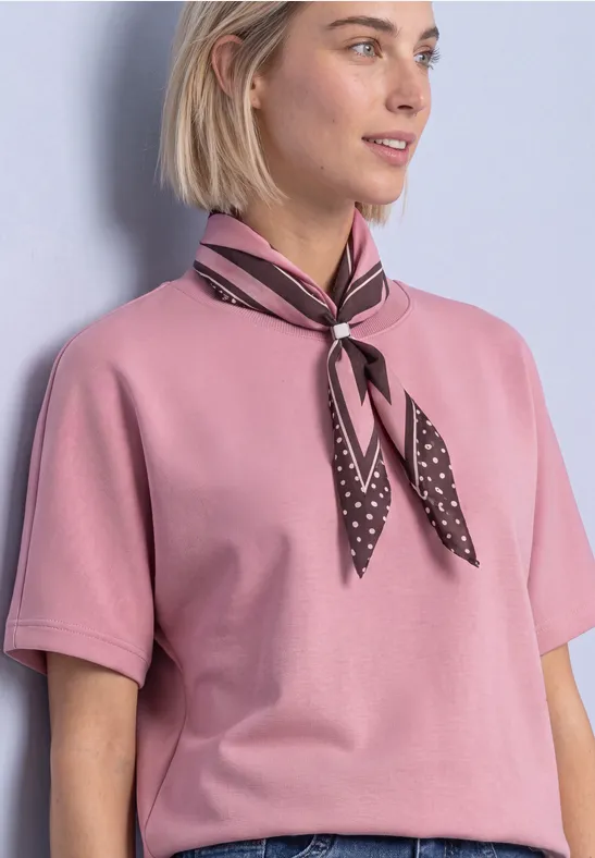 Femme en t-shirt rose à manches courtes et foulard à pois noué autour du cou devant un mur clair.