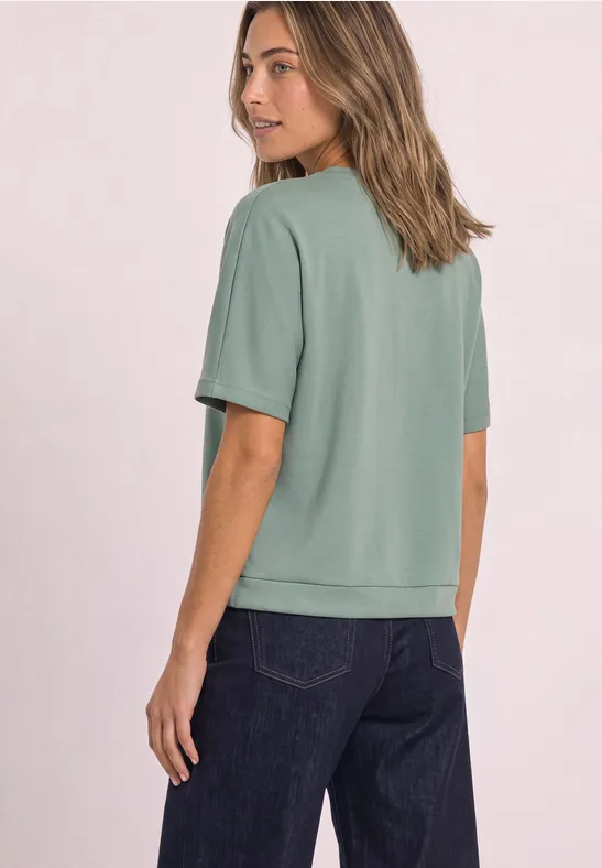 Vrouw van achteren in een loszittend groen T-shirt en donkere spijkerbroek.
