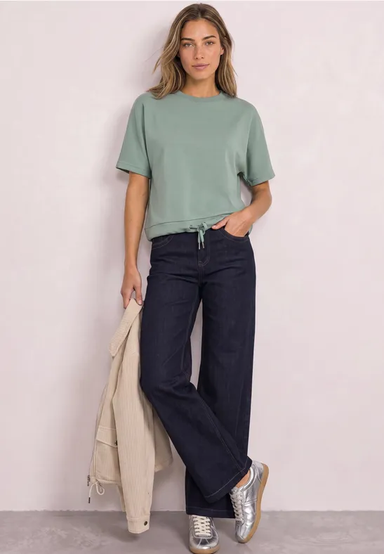 Vrouw in groen T-shirt, donkere wijdvallende jeans en sneakers, met een beige jas in haar hand.