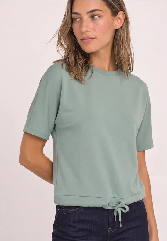 Een jonge vrouw draagt een saliegroene trui met korte mouwen en aantrekkoord in de zoom, met donkerblauwe spijkerbroek.