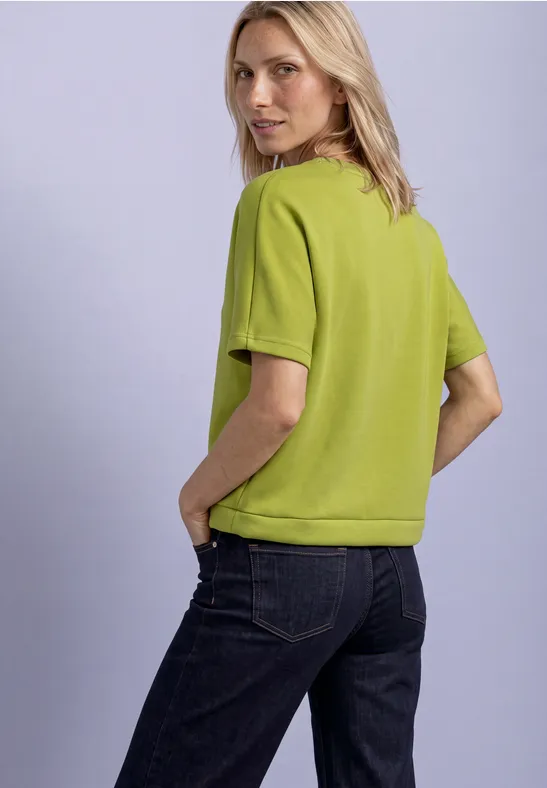 Frau in grüner Kurzarmbluse und dunklen Jeans schaut über ihre Schulter, Hände in den Taschen.