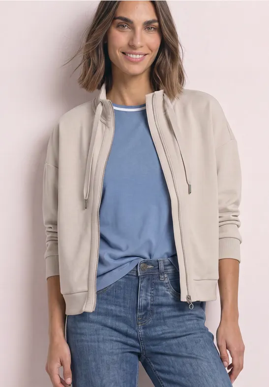 Frau trägt beige Jacke, blaues T-Shirt und Jeans, lächelt vor hellem Hintergrund.