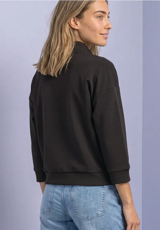 Frau von hinten in schwarzer Jacke und blauen Jeans, leicht zur Seite schauend vor hellem Hintergrund.