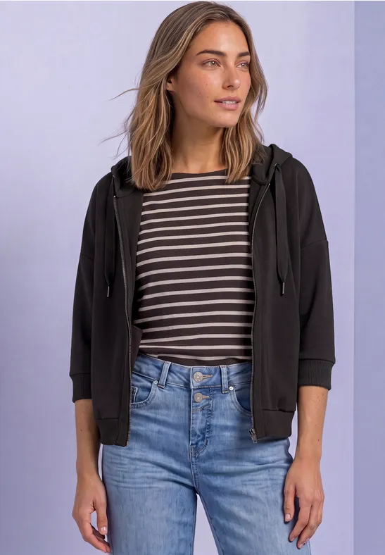 Frau mit gestreiftem Shirt, schwarzer Kapuzenjacke und Jeans steht vor hellem Hintergrund.