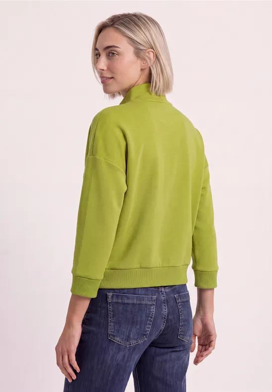 Femme vue de dos portant un pull vert et un jean sur fond clair.