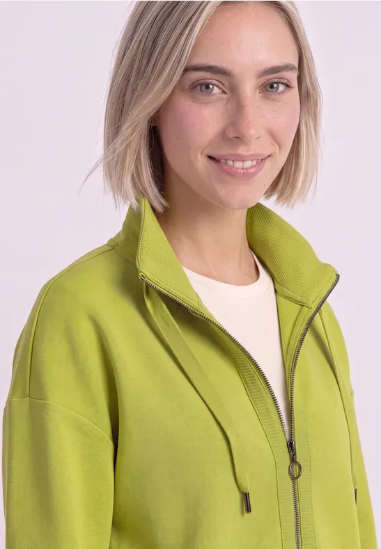Femme aux cheveux blonds courts souriante, portant une veste zippée vert clair et un t-shirt blanc.